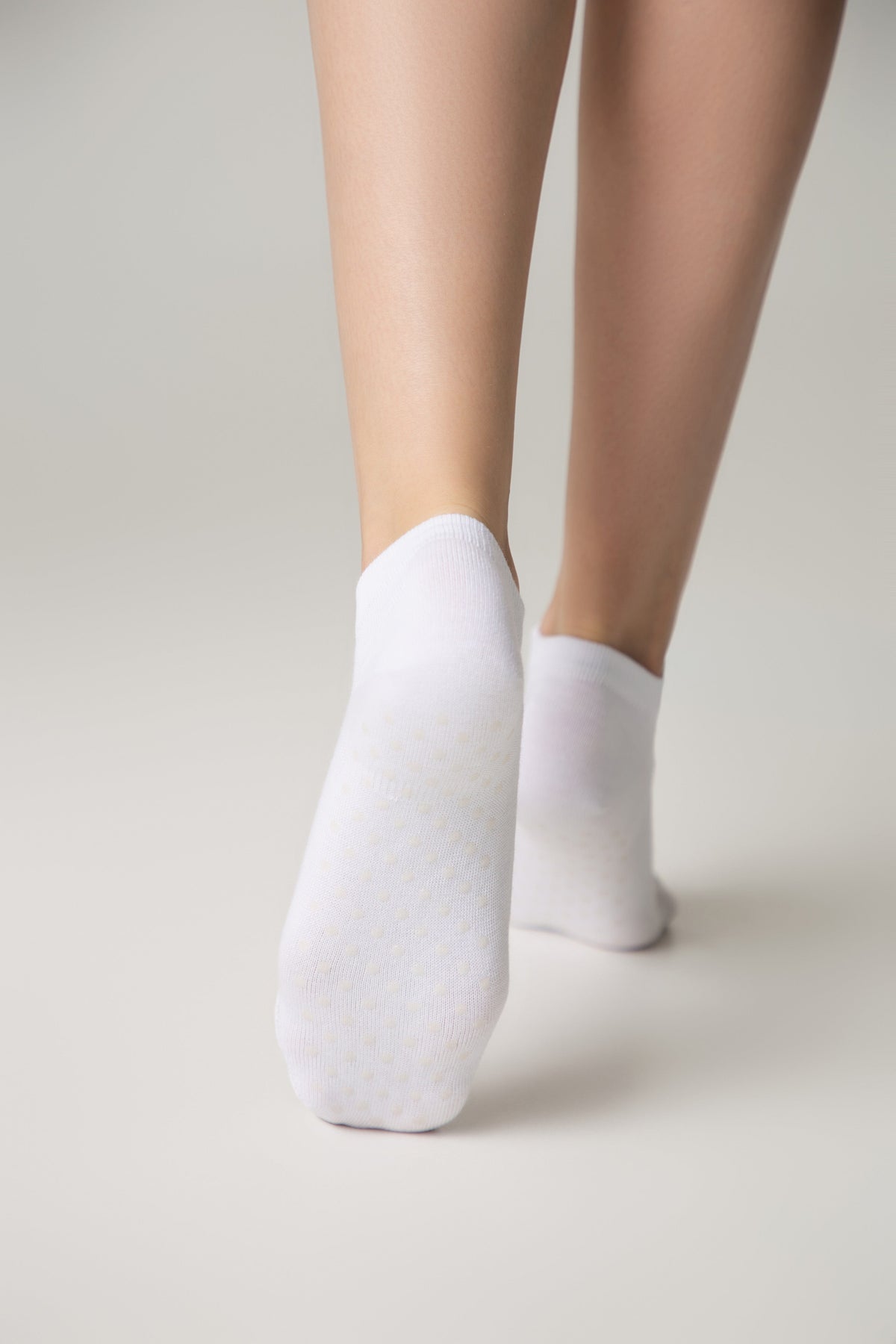 A pair of white ankle socks worn on feet with visible silicone grip dots on the sole, set against a plain light grey background.