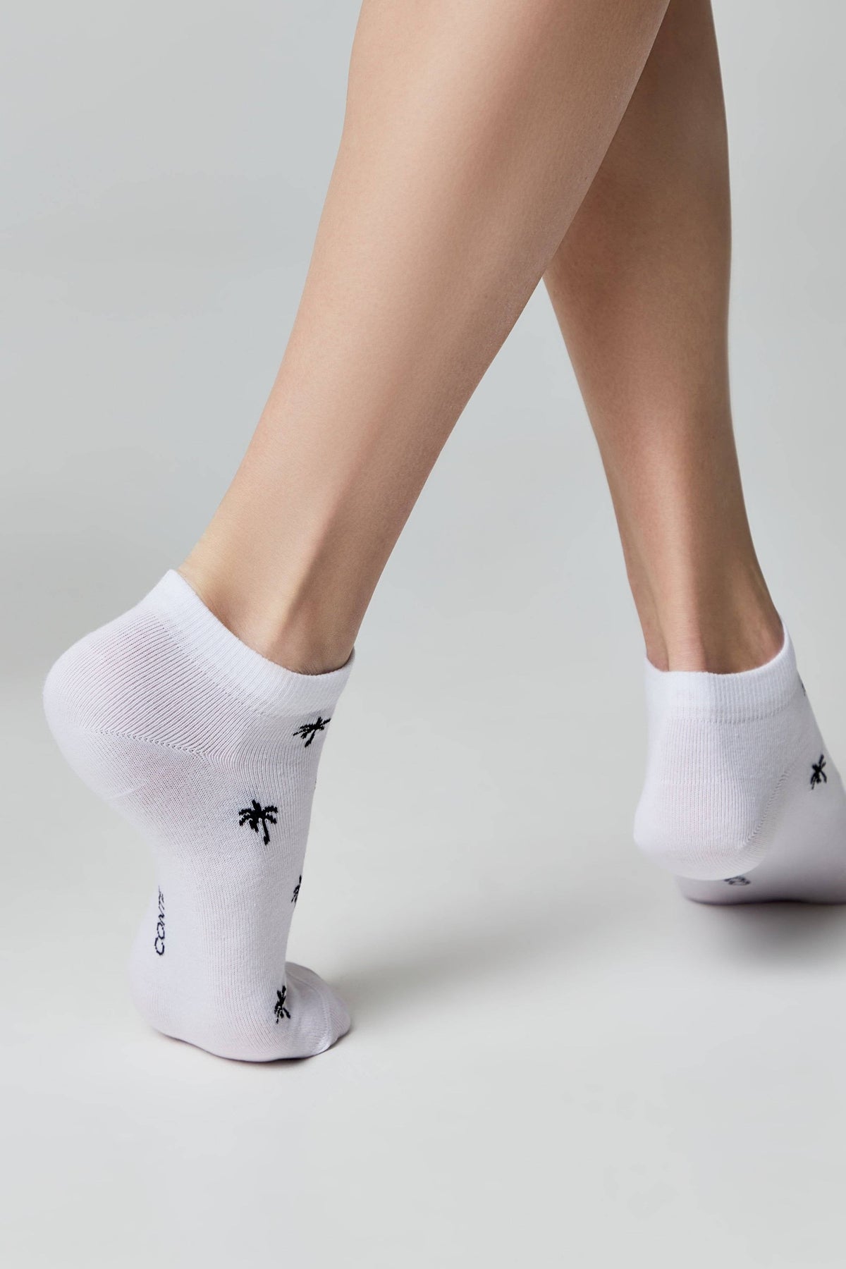A pair of white ankle socks patterned with small black palm tree icons, worn on the feet of a person standing against a plain light gray background.