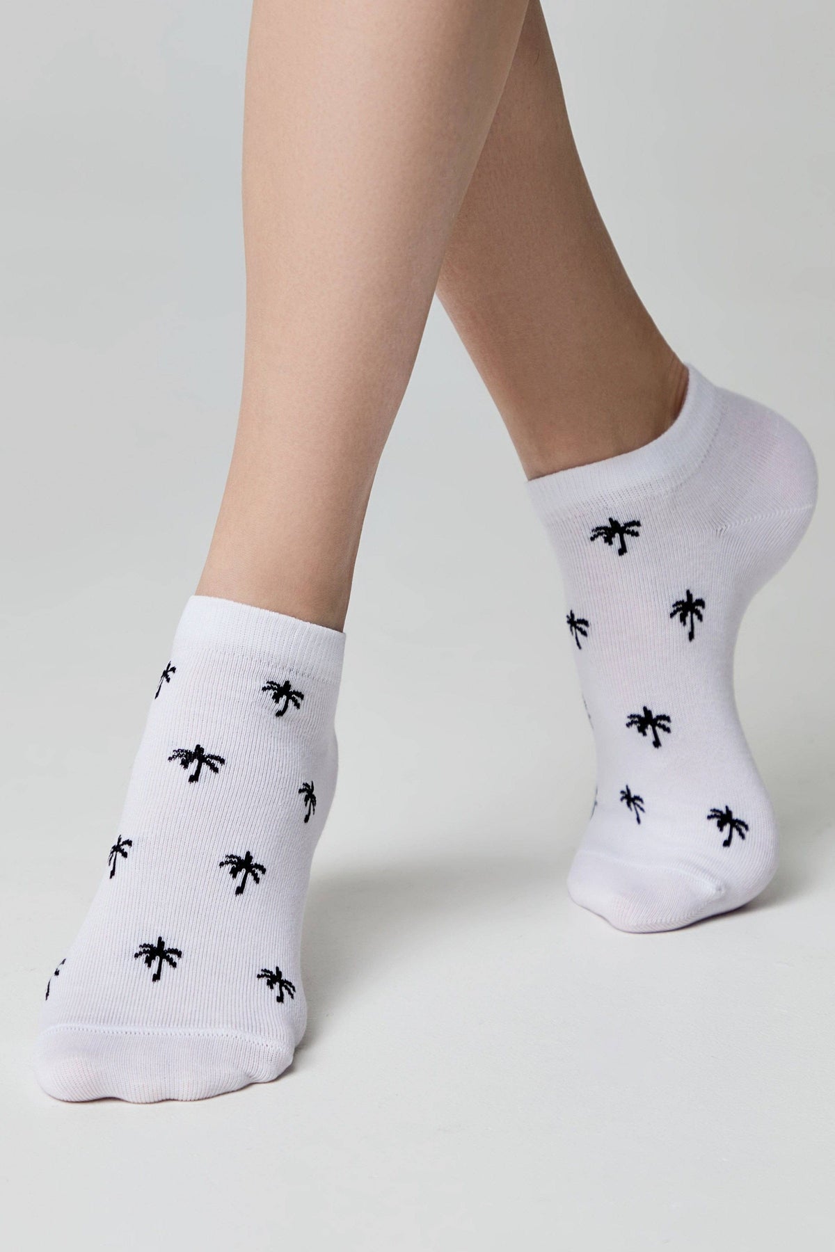 White ankle socks with a repeating small black palm tree pattern worn on feet against a light grey background.
