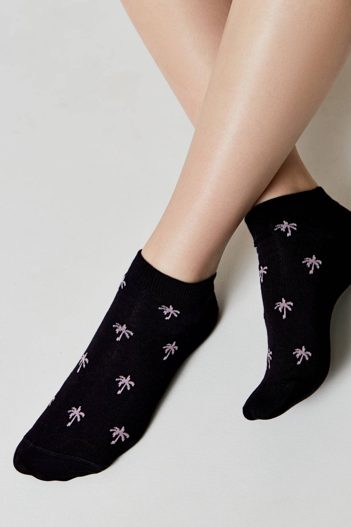 A pair of black ankle socks decorated with a small, repeating pattern of light pink palm trees worn on a person's feet against a white background.