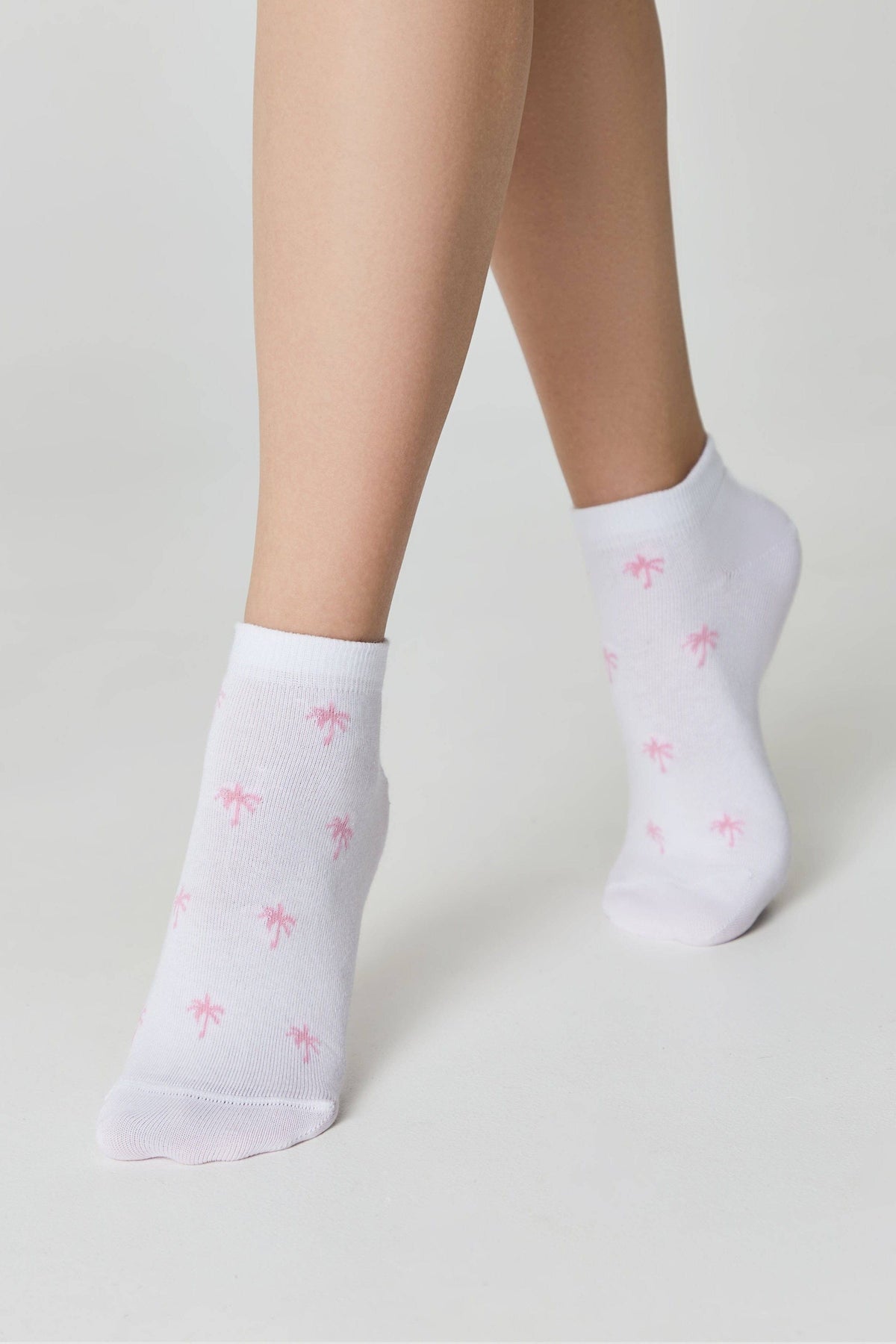 A pair of white ankle socks featuring a small pink palm tree pattern, modeled on a person's legs against a plain light background.