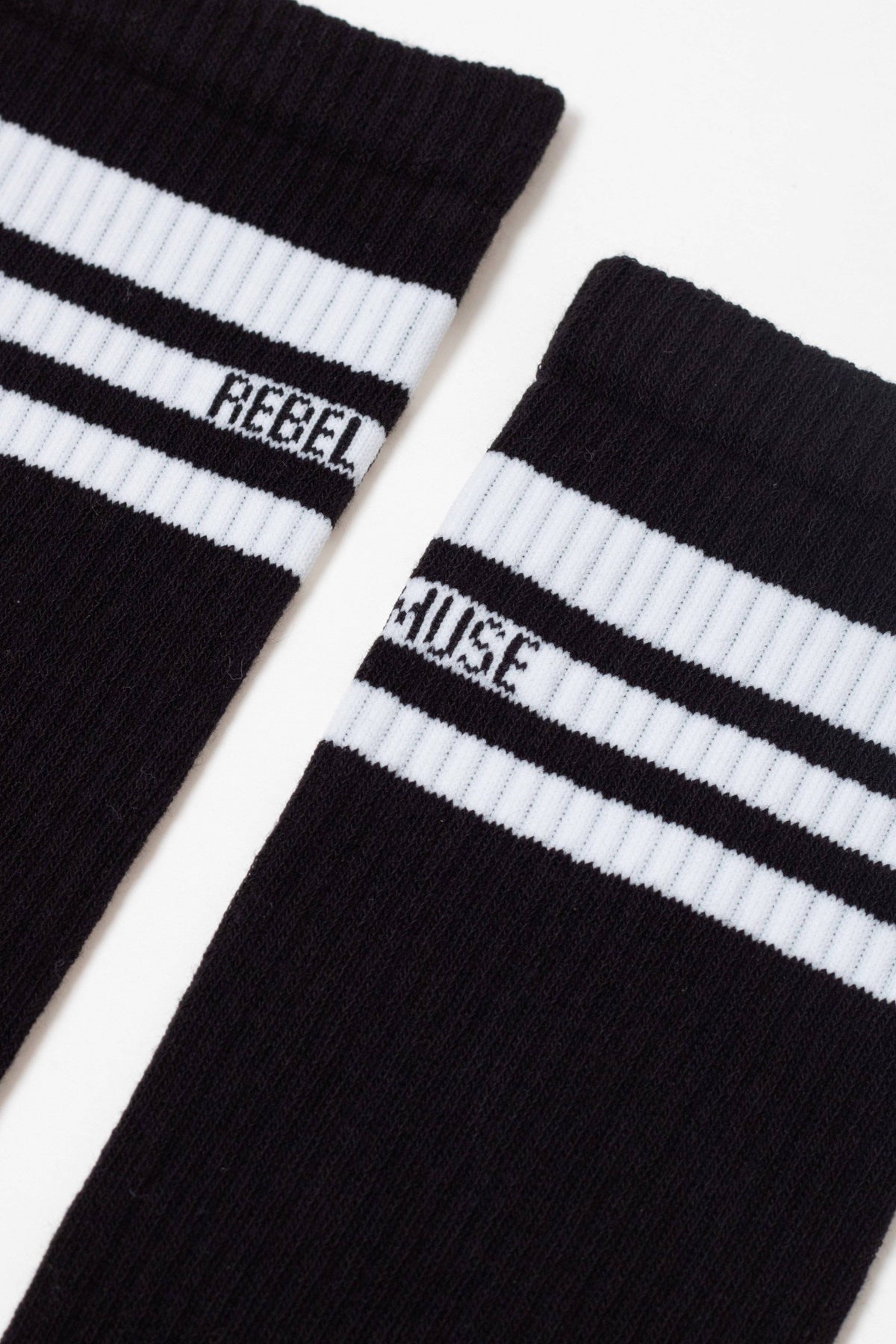 Two black crew socks are laid side-by-side, each featuring two white stripes around the cuff with the word REBEL printed on the left sock and MUSE printed on the right sock.