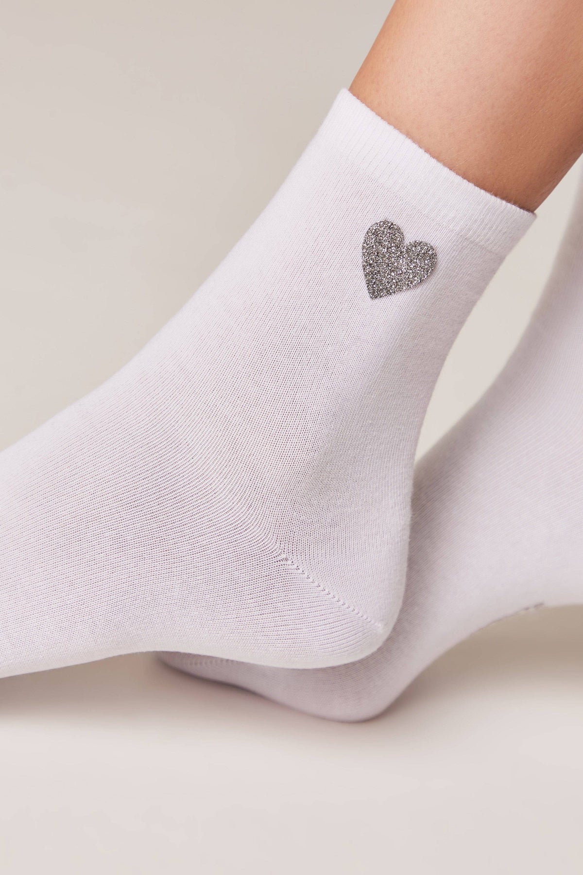White ankle socks featuring a small, glittery silver heart detail on the side, worn on a person's feet against a light background.