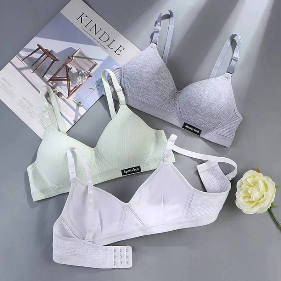 Three soft, wireless bras in grey, mint green, and white are laid out on a flat surface next to a Kindle magazine and a decorative white rose.