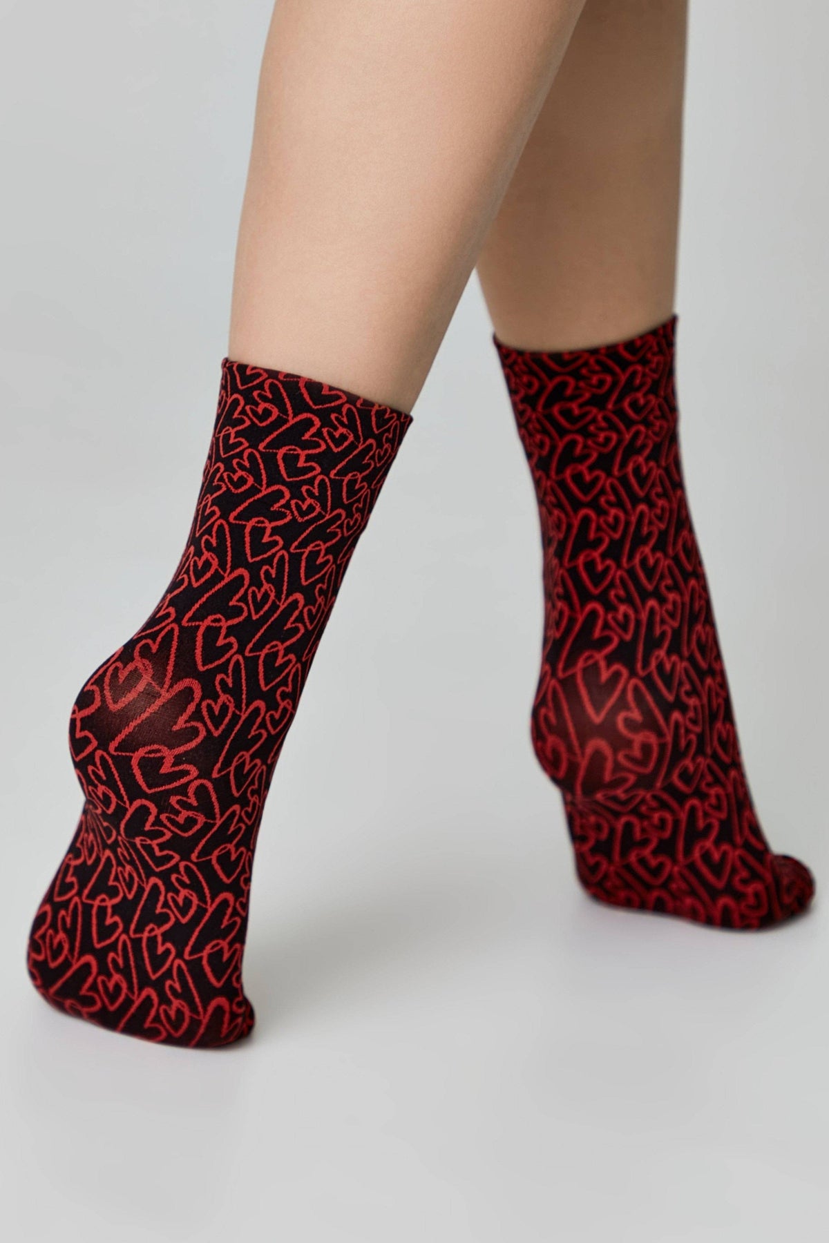 Ankle socks featuring a repeating pattern of red outlined hearts on a black background worn on a person's lower legs against a neutral backdrop.