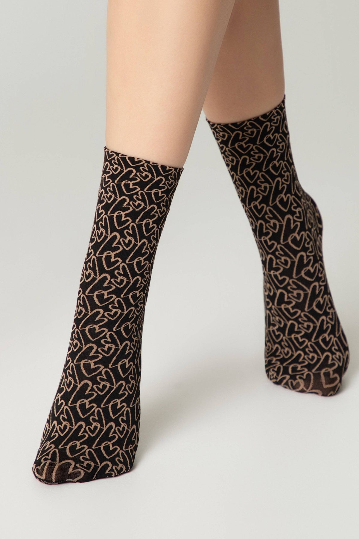 A pair of sheer black crew socks featuring an all-over pattern of thin, light brown outline hearts worn on a person's lower legs against a plain light gray background.