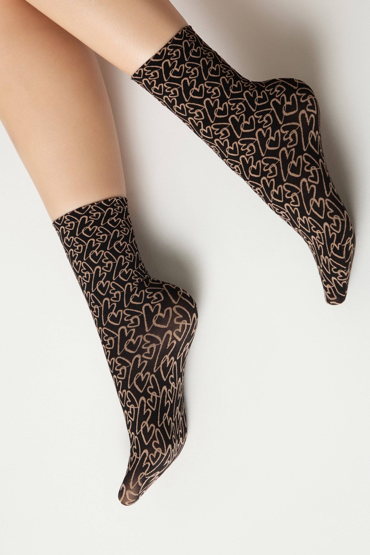 A pair of sheer black crew socks decorated with a repeating pattern of outlined beige hearts worn on a person's legs against a plain white background.