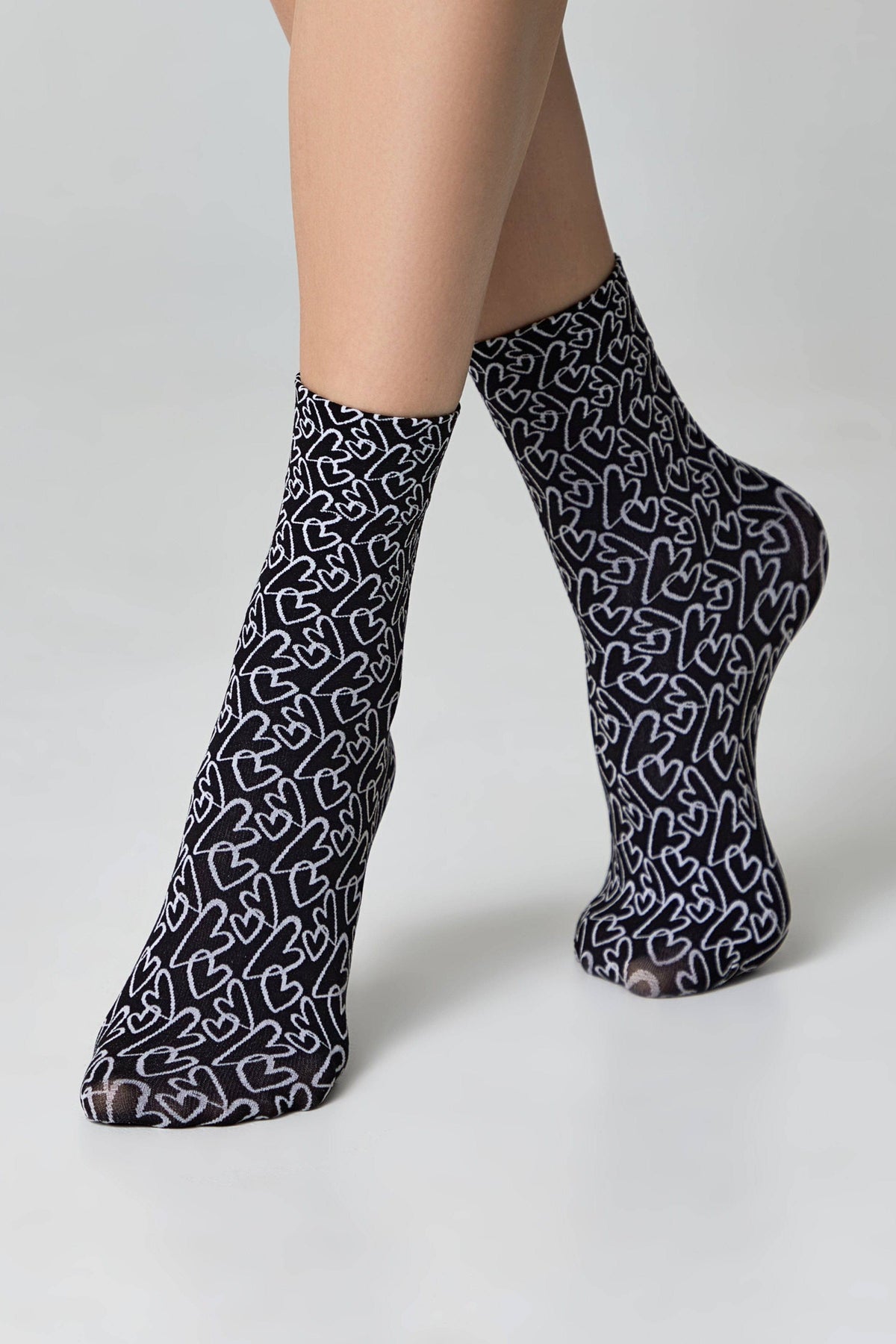 Sheer black ankle socks patterned with repeating white outlined hearts worn on a person's feet against a light grey background.