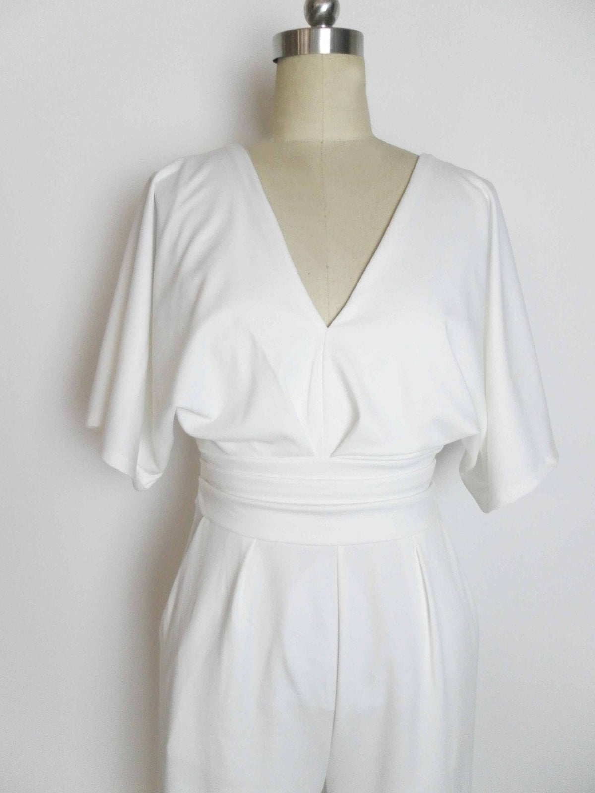 A white jumpsuit on a mannequin features a deep V-neckline, loose elbow-length sleeves, and a wide, gathered waistband.