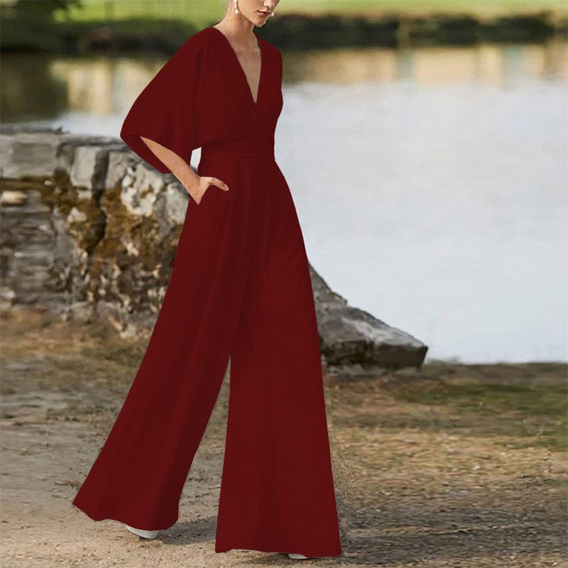 A woman stands outdoors wearing a deep red, long-sleeved jumpsuit with a deep v-neck and wide-leg trousers.
