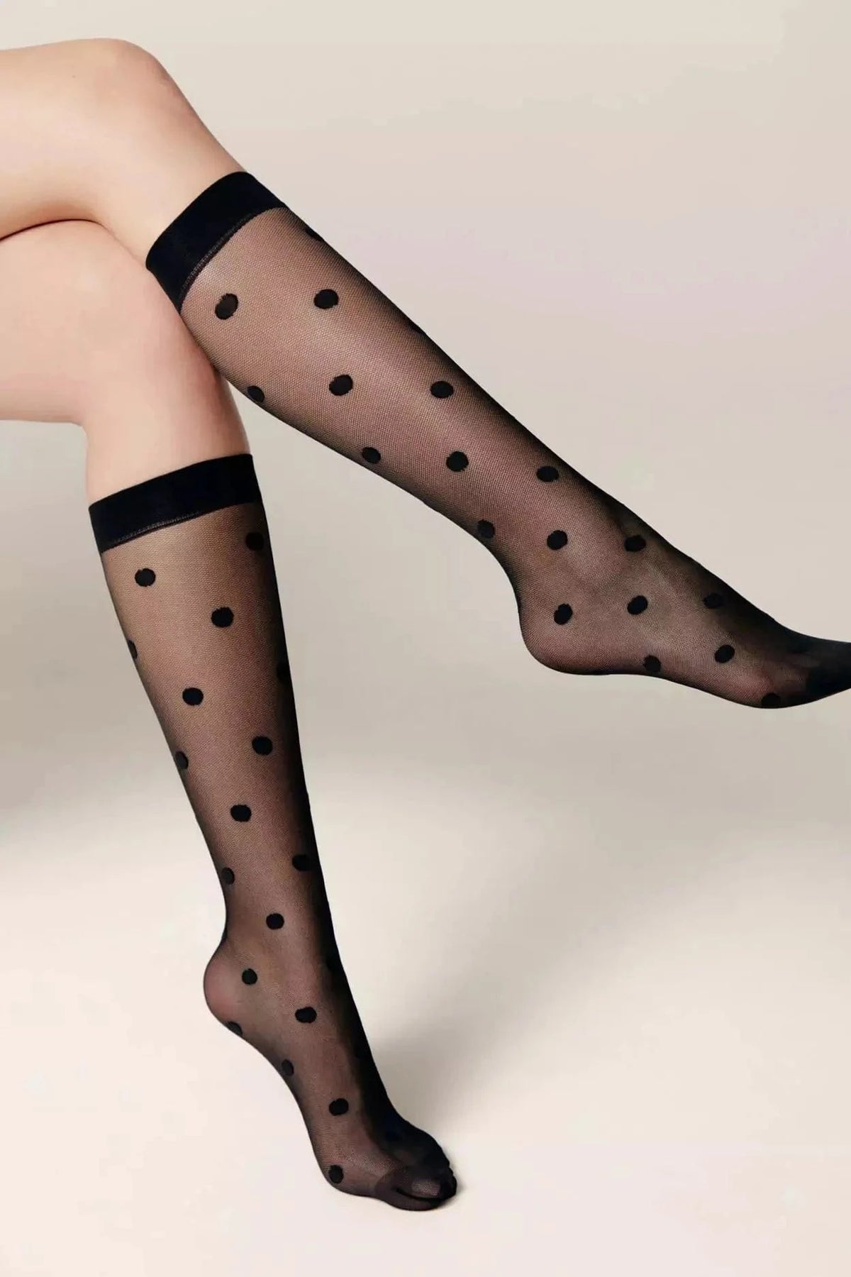 Sheer black knee-high stockings featuring a repeating black polka dot pattern and a solid black ribbed top band worn on a person's legs.