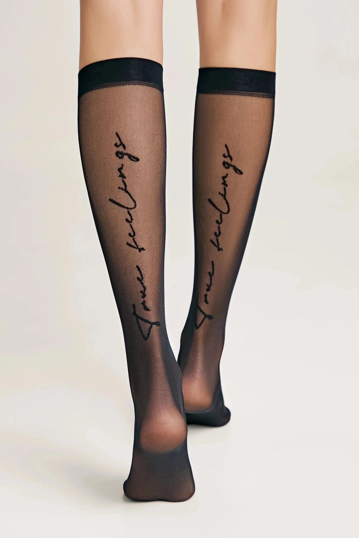 A pair of sheer black knee-high stockings featuring the words true feelings written in a cursive black font running vertically down the back of each calf.