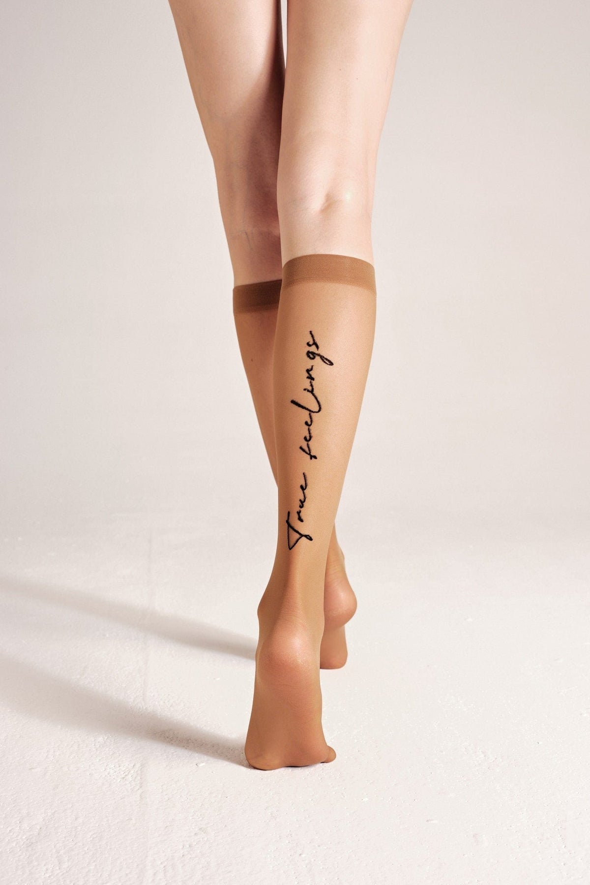 A pair of sheer tan knee-high stockings worn on a person's legs, featuring the script text True feelings printed vertically in black on the calf.