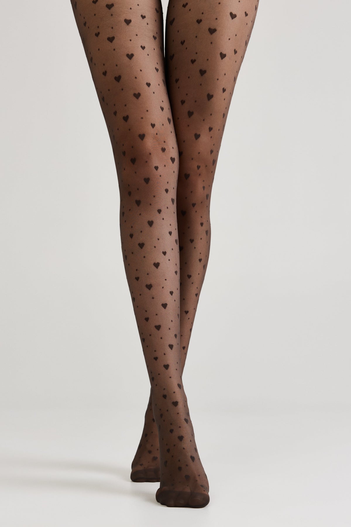 Sheer black tights patterned with small dark hearts worn on a person's legs against a plain light gray background.