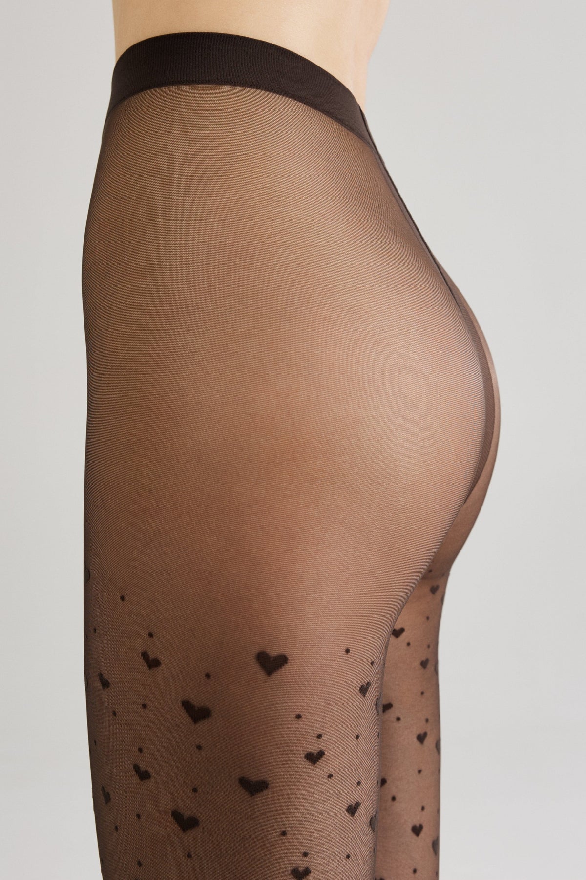 Sheer black tights featuring a repeating pattern of small dark hearts and scattered dots worn on a person's lower torso and legs against a light grey background.