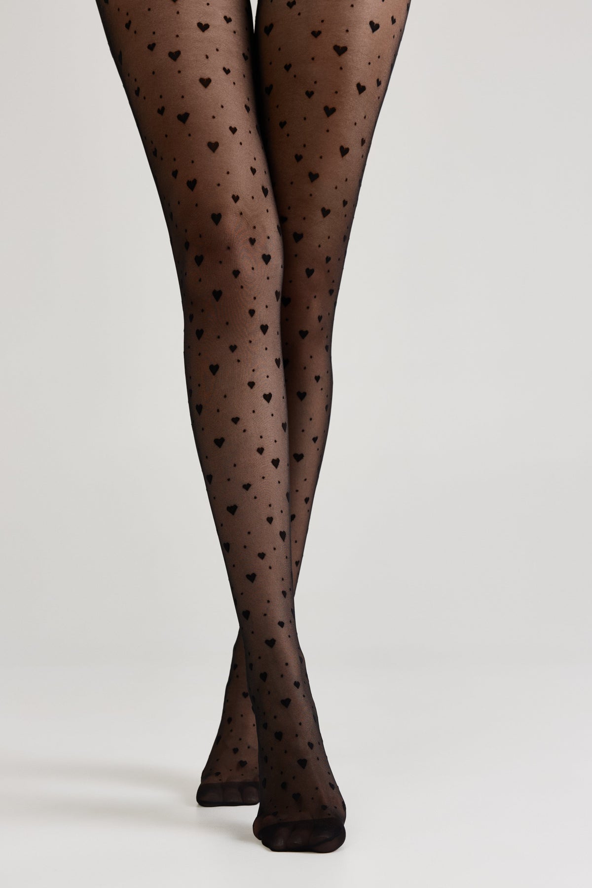 Sheer black tights decorated with a recurring small heart pattern worn on legs against a plain light gray background.