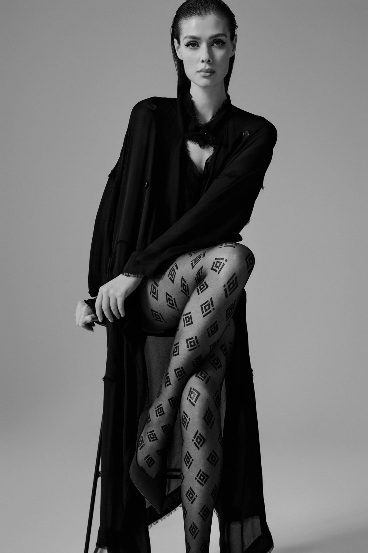 Patterned tights in black featuring a repeating geometric diamond logo print worn by a person dressed in an oversized black coat.