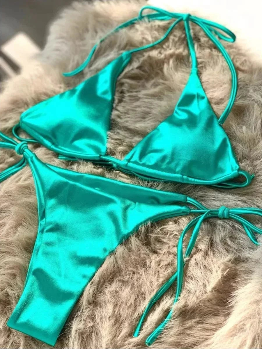 A vibrant turquoise satin bikini set consisting of a triangle top and matching side-tie thong bottoms, displayed on a fluffy beige faux fur surface.