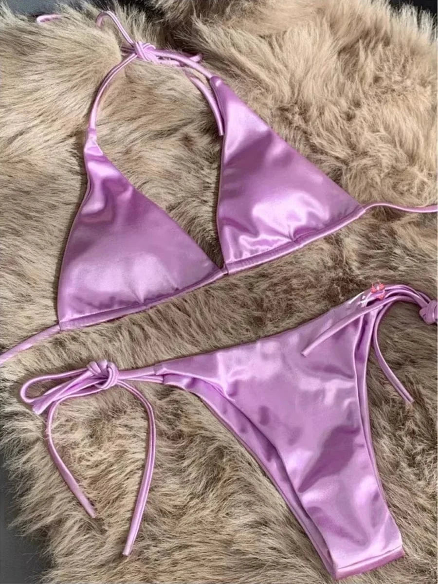 A light purple satin two-piece bikini set with a triangle top and side-tie bottoms displayed on a fuzzy textured surface.