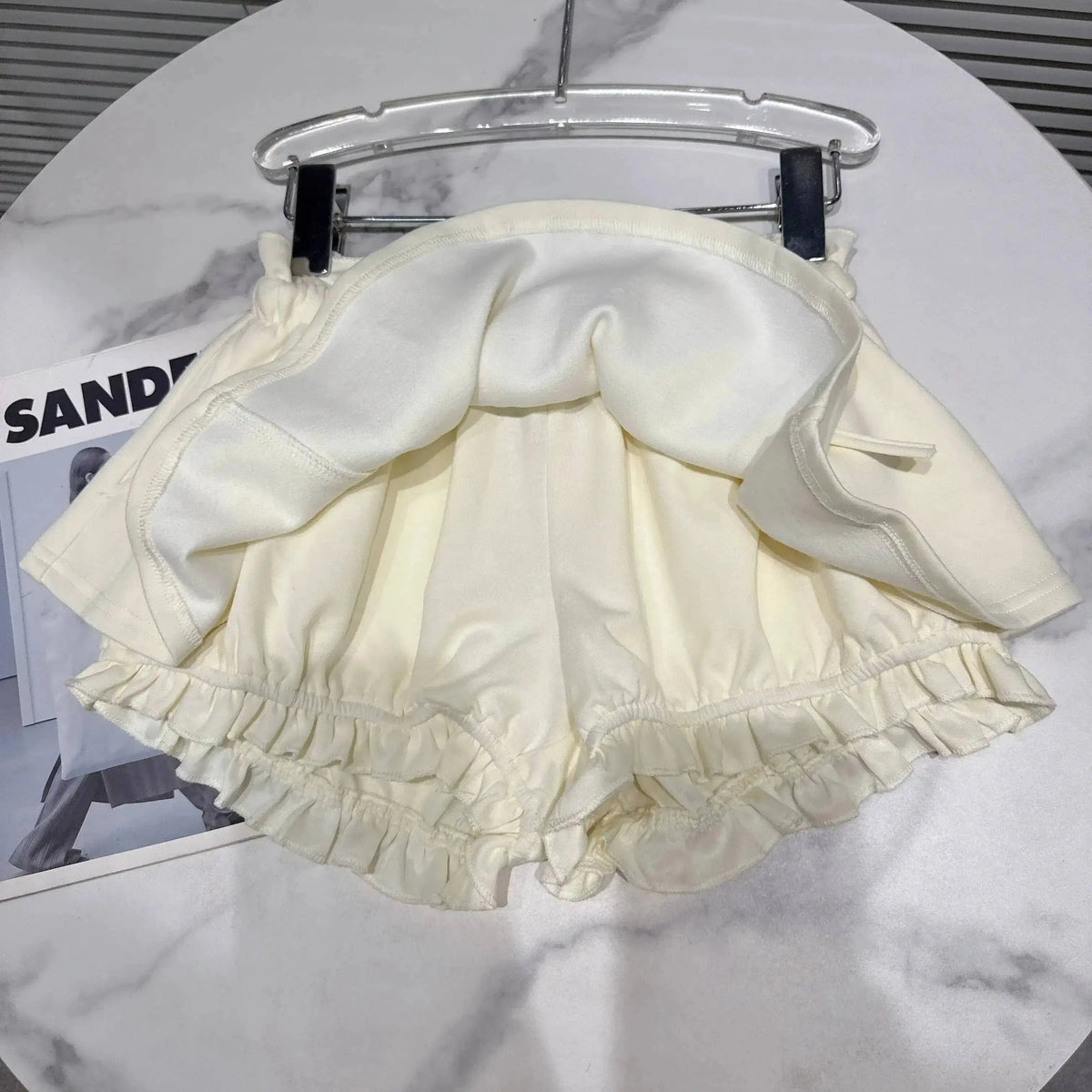 A cream-colored skirt with integrated shorts featuring a ruffled hemline, suspended on a clear clothes hanger against a marbled surface.
