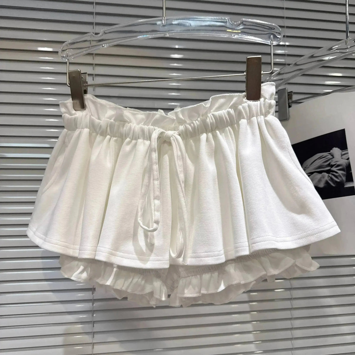 A white tiered mini skirt with an elasticated drawstring waistband hanging on a clear plastic hanger against a horizontal slat blind background.