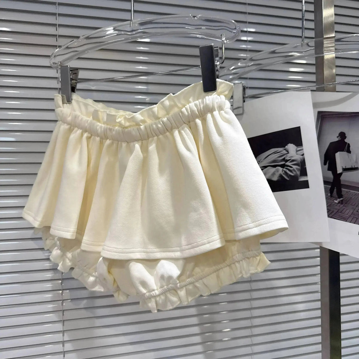 A pair of cream-colored, multi-layered ruffled shorts with an elasticized waistband hanging from a clear plastic hanger against a slatted window blind.