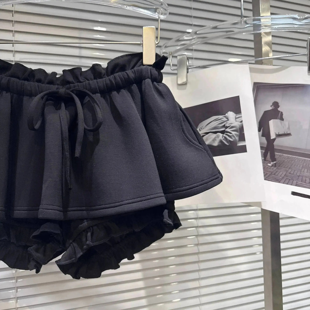 Black ruffled shorts with an elasticated drawstring waistband hanging on a display rack in front of white blinds.