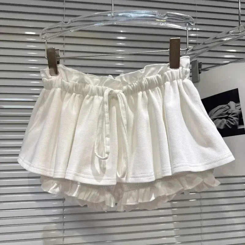 A white, tiered ruffled mini skirt or shorts with an elasticated waistband and a drawstring closure, displayed on a hanger against a slatted blind background.