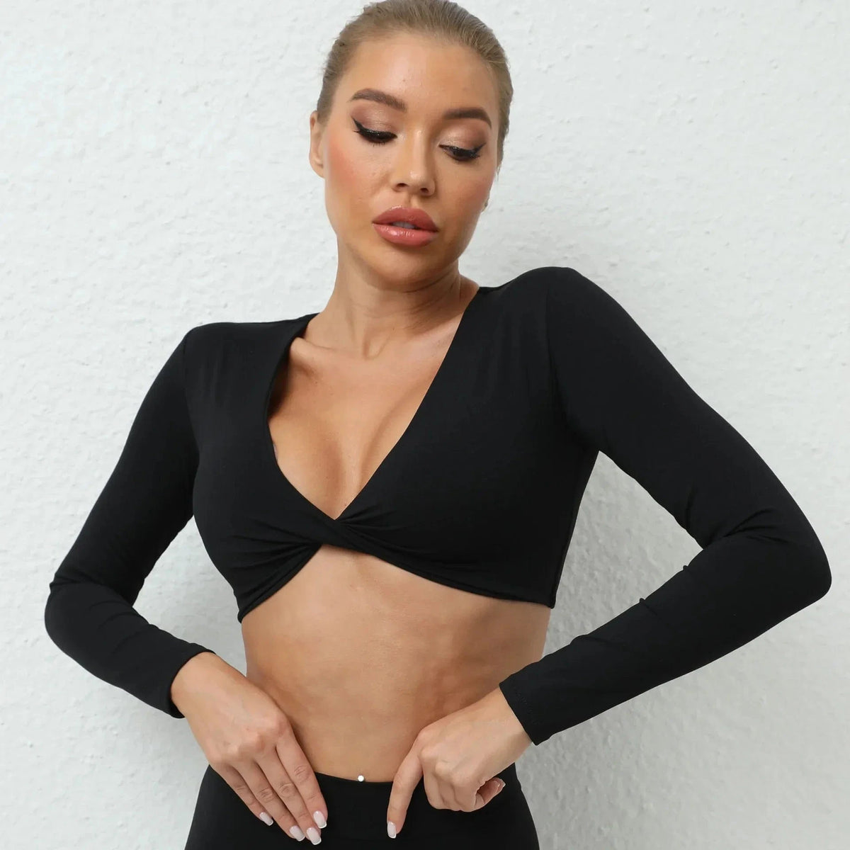 A woman wearing a black long-sleeved cropped top with a twisted detail at the center of the V-neckline against a white textured background.