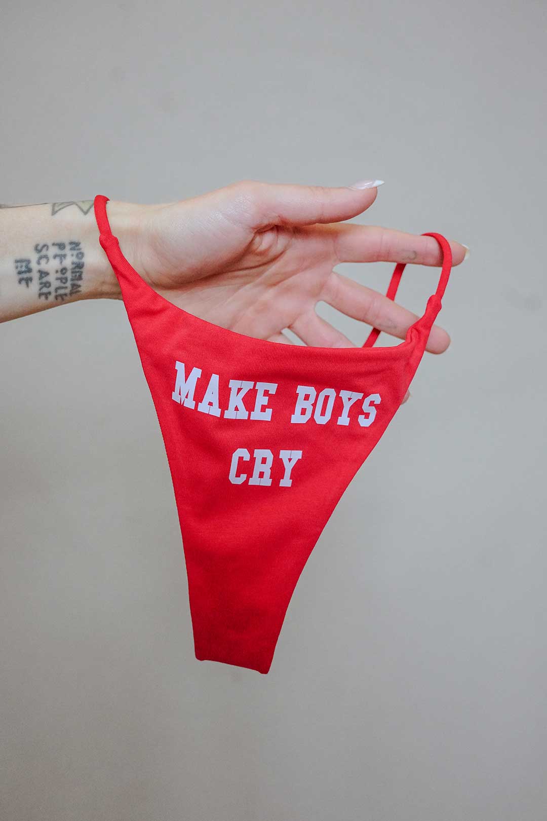 A red thong swimsuit bottom featuring the white text "MAKE BOYS CRY" is held up by a hand against a plain background.