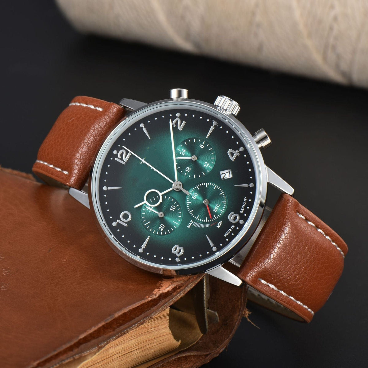 A round watch with a green sunburst dial featuring three sub-dials, a date window, and silver indices on a brown leather strap resting against a brown leather notebook.