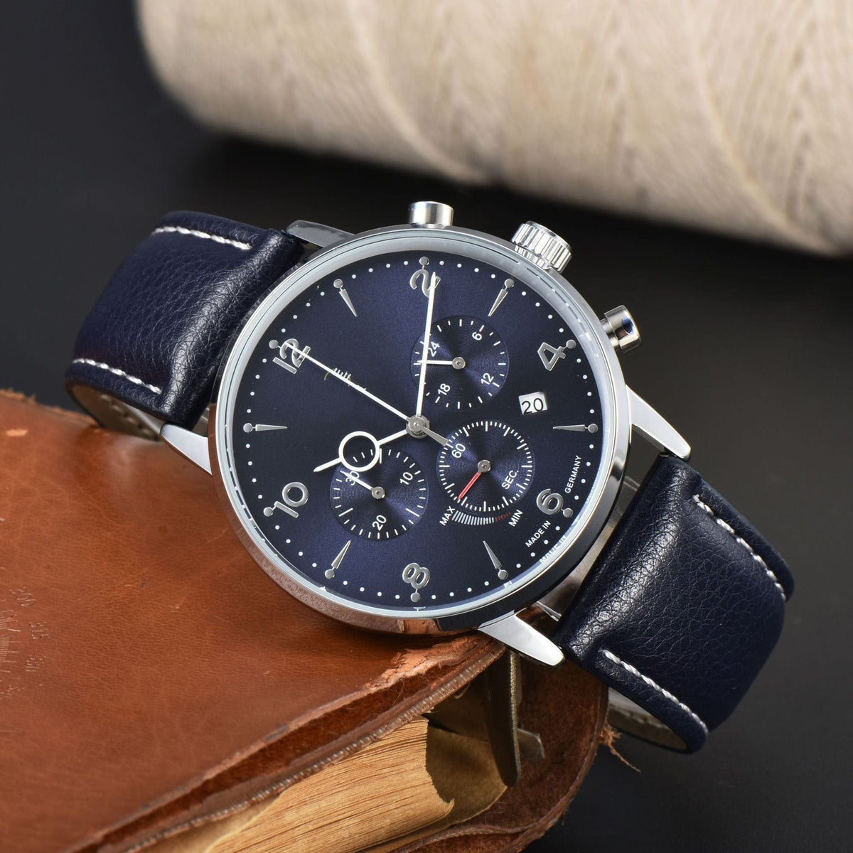 A round silver-toned chronograph watch with a deep blue dial, white numeral hour markers, and a blue leather strap featuring white contrast stitching, resting on a brown leather-covered book.