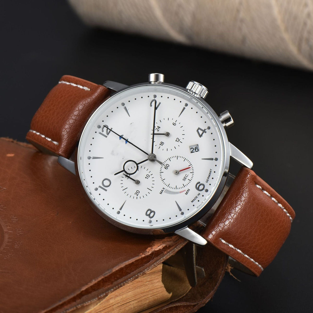 A round chronograph watch with a white dial, brown leather strap, and silver-tone case resting on a leather-bound book against a dark background.