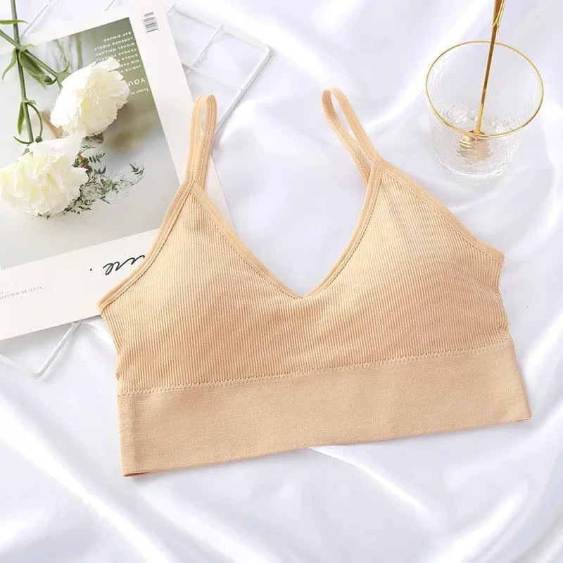 A beige ribbed V-neck bralette with spaghetti straps is displayed on a white satin fabric surface next to a white card with flowers and a clear glass.