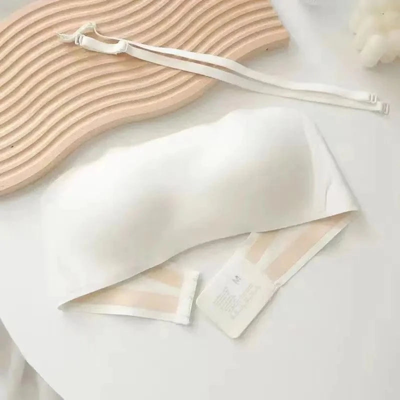 A strapless white bandeau bra lies on a white table next to a pair of detachable white bra straps and a decorative wavy wooden board.