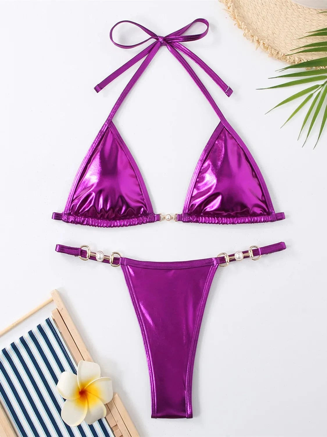 A metallic purple two-piece bikini set featuring a triangle top and high-cut bottoms with gold-toned ring details and pearl accents, laid flat on a white surface with a striped chair and a tropical flower.
