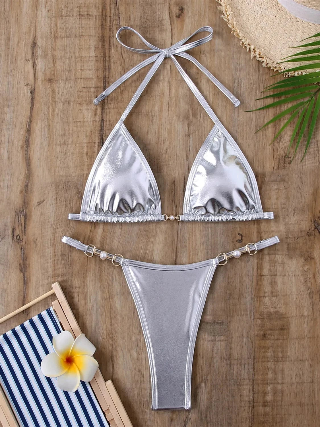 A metallic silver two-piece bikini set featuring triangular cups with a pearl detail at the center and matching bottoms with pearl-accented ring side connectors, laid flat on a wooden surface.