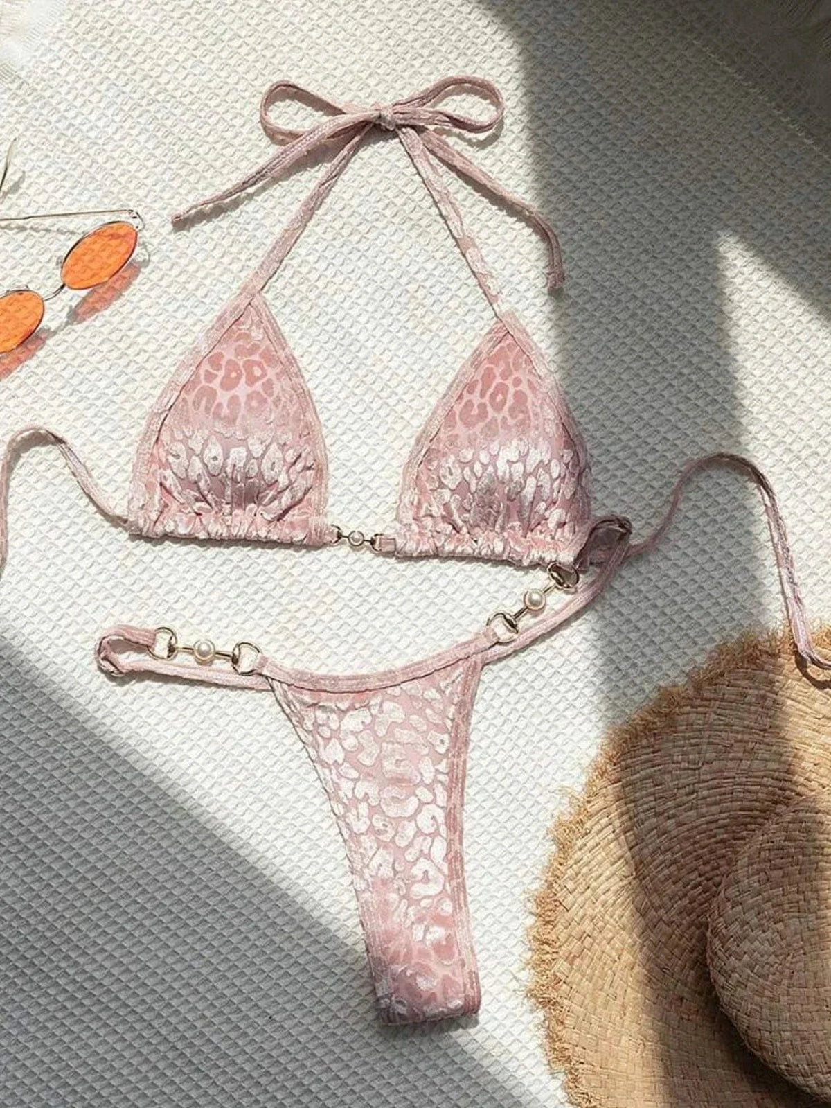 A light pink leopard print bikini set featuring a triangle top and high-cut bottoms, both accented with gold-toned hardware and faux pearl beads, laid out on a textured surface next to orange sunglasses and a straw hat.