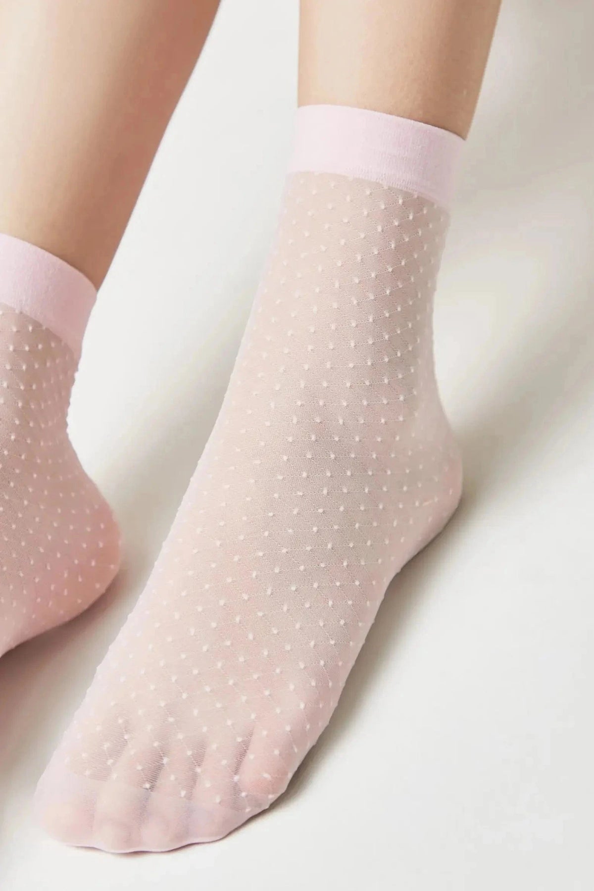 Sheer pale pink ankle socks featuring a delicate swiss dot pattern and solid light pink ribbed cuffs worn on a person's feet.