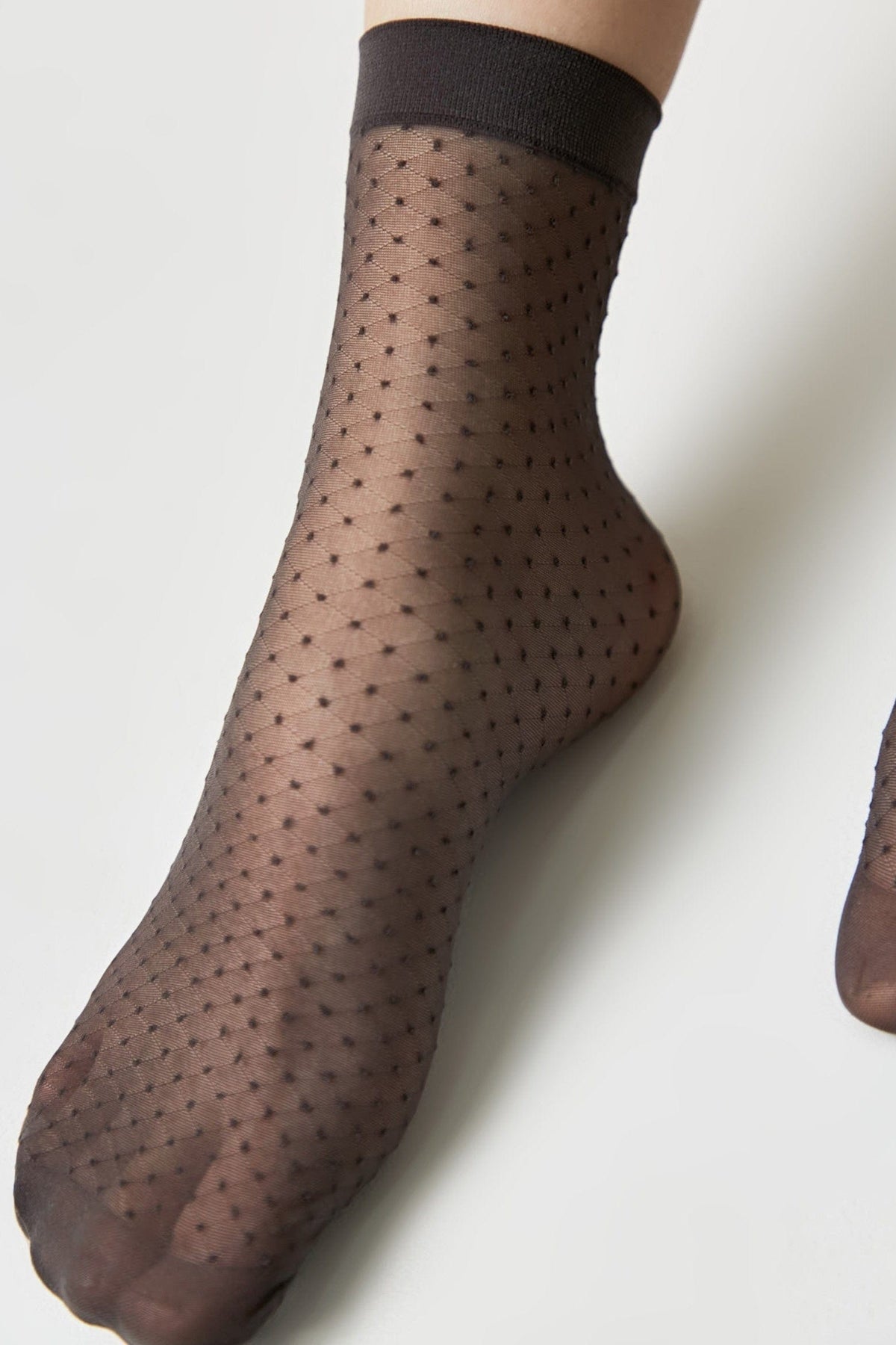A close-up of a foot wearing a sheer black ankle sock with a subtle diamond pattern and small dots on a plain white background.