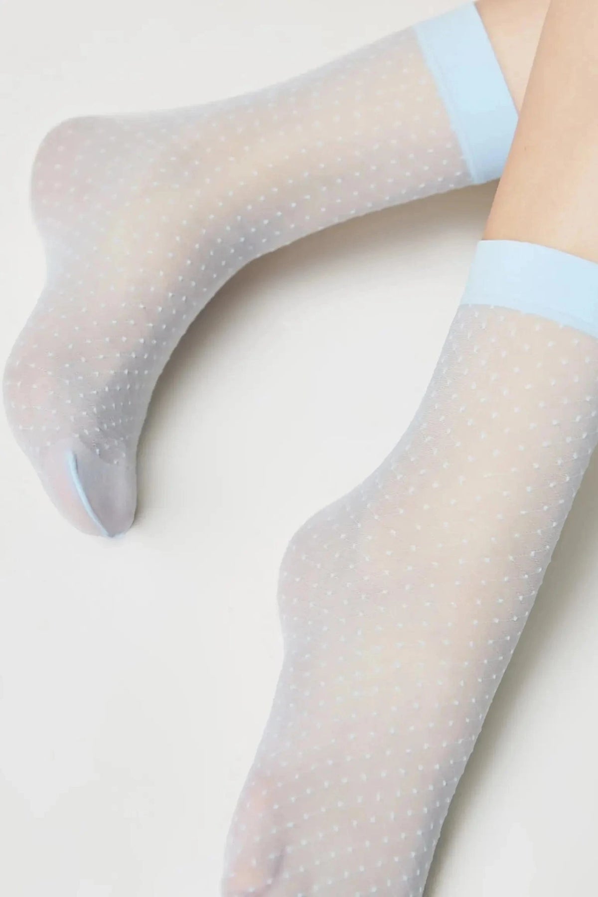 Sheer white ankle socks featuring a subtle white polka dot pattern and light blue trim at the cuff, shown worn on a pair of feet.