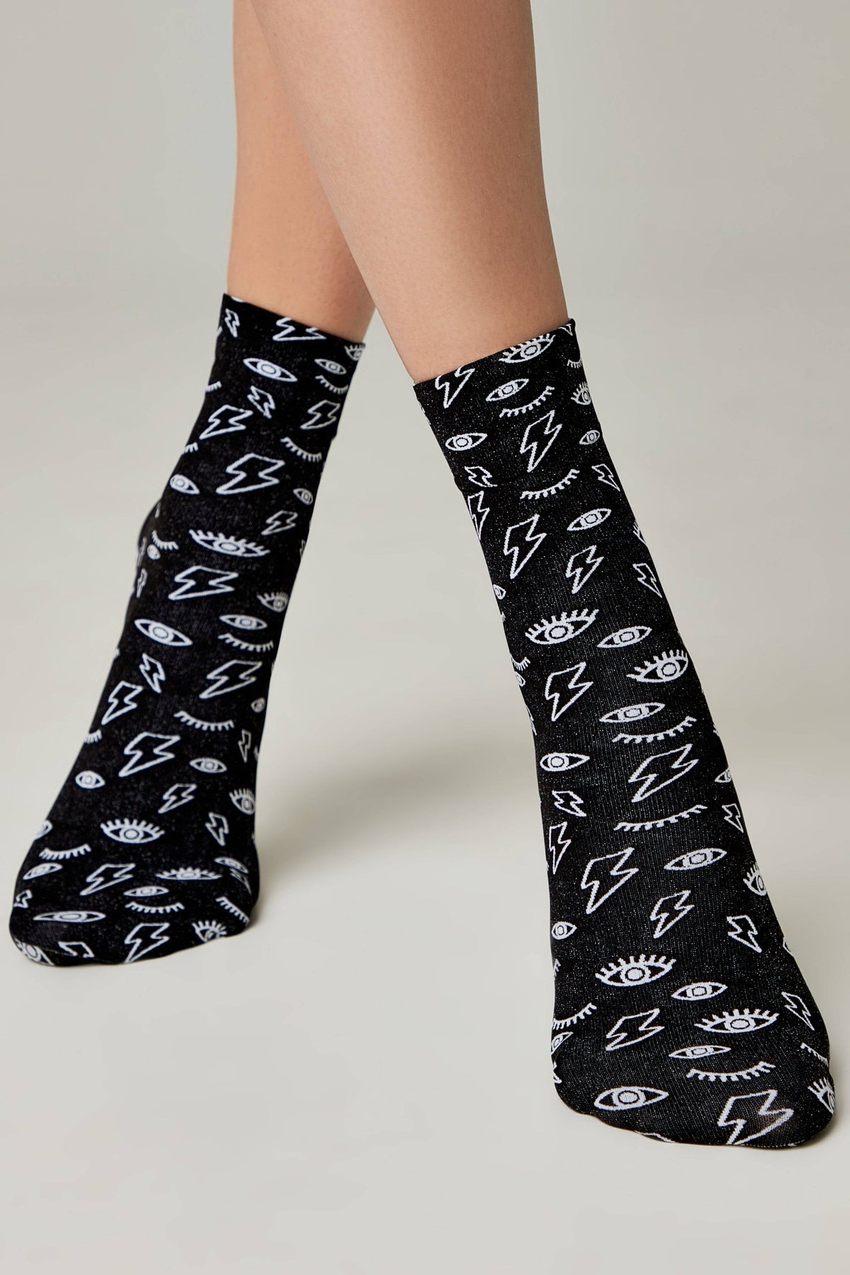 A pair of black crew-length socks features a repeating white pattern of stylized lightning bolts and eyes with long eyelashes worn on a person's feet.