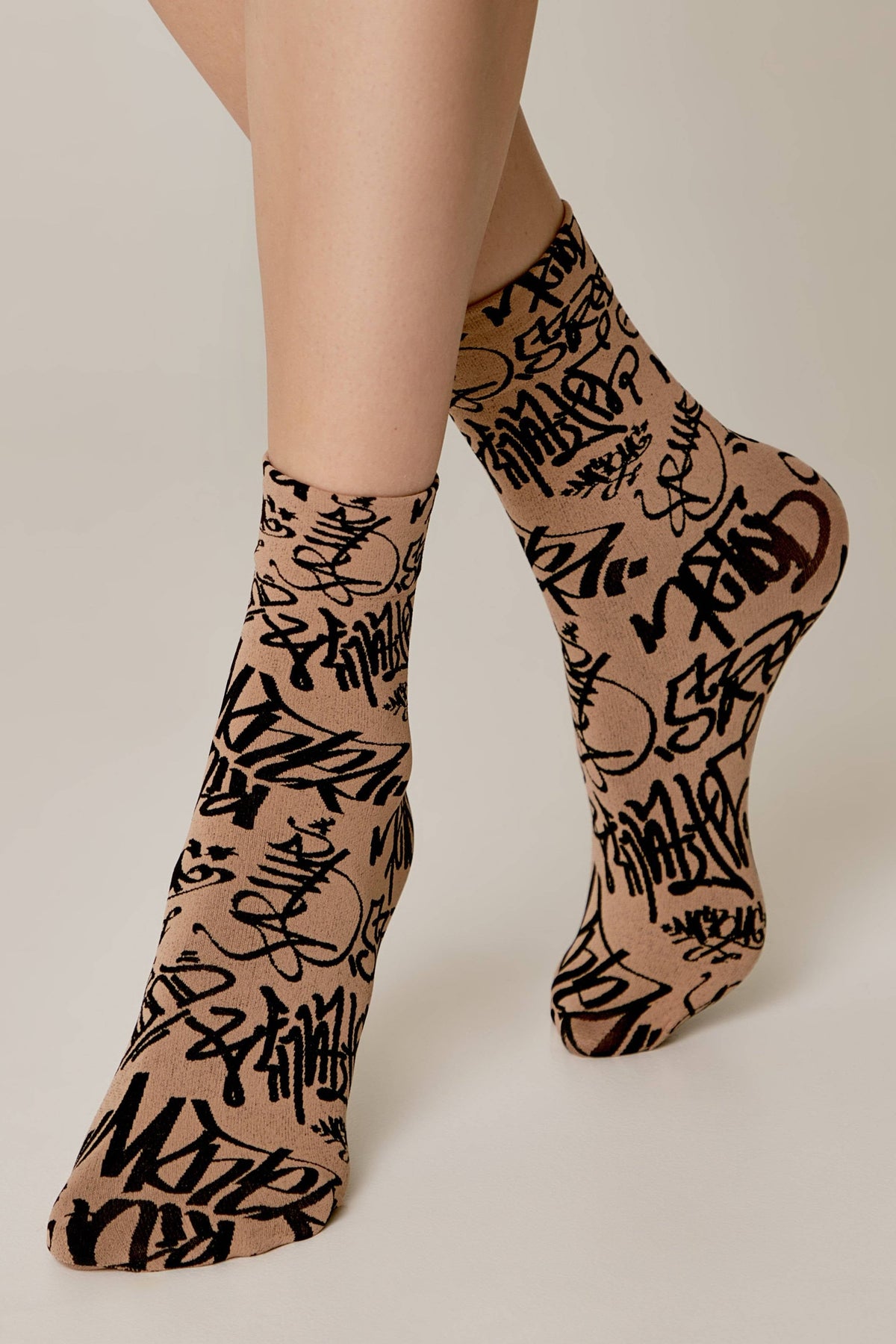 A pair of tan-colored ankle socks with a black graffiti-style text print worn on a person's feet against a light background.
