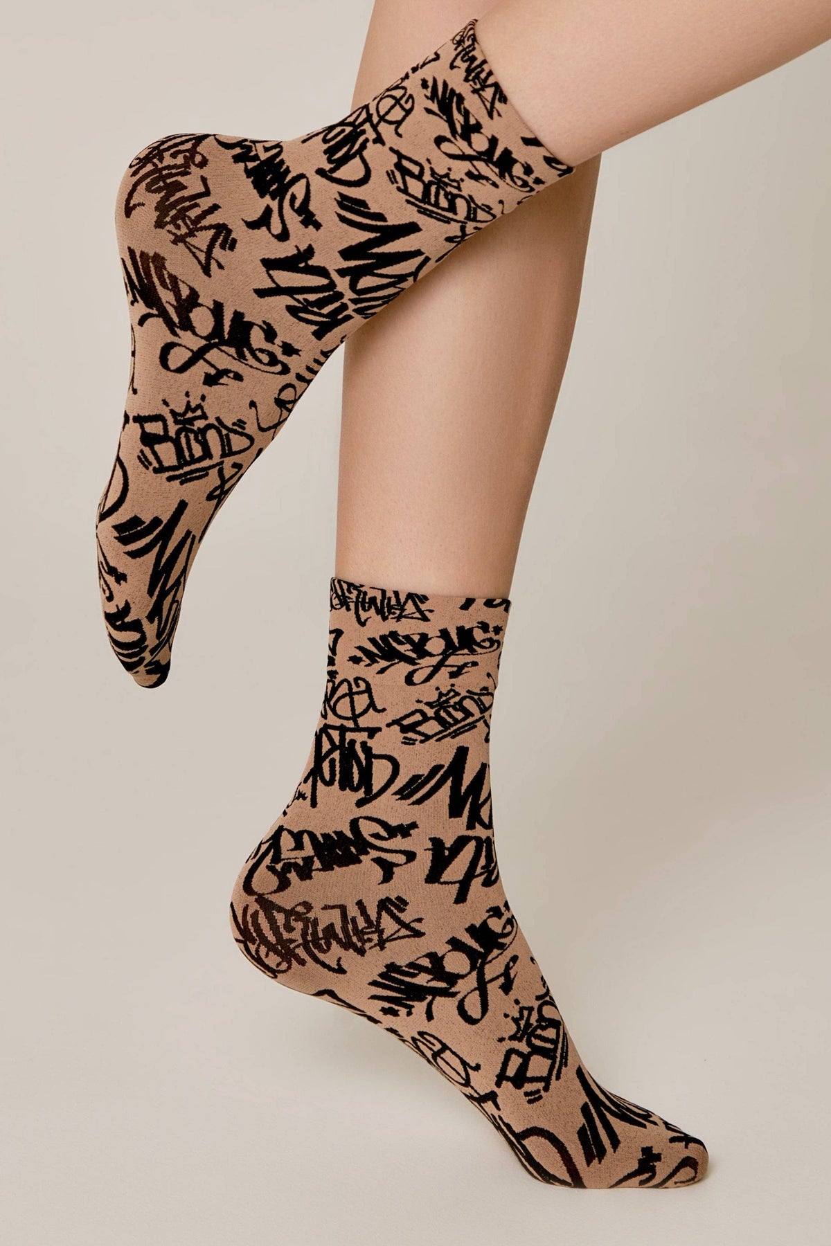 A pair of tan ankle socks featuring a black graffiti-style pattern worn on a person's feet.