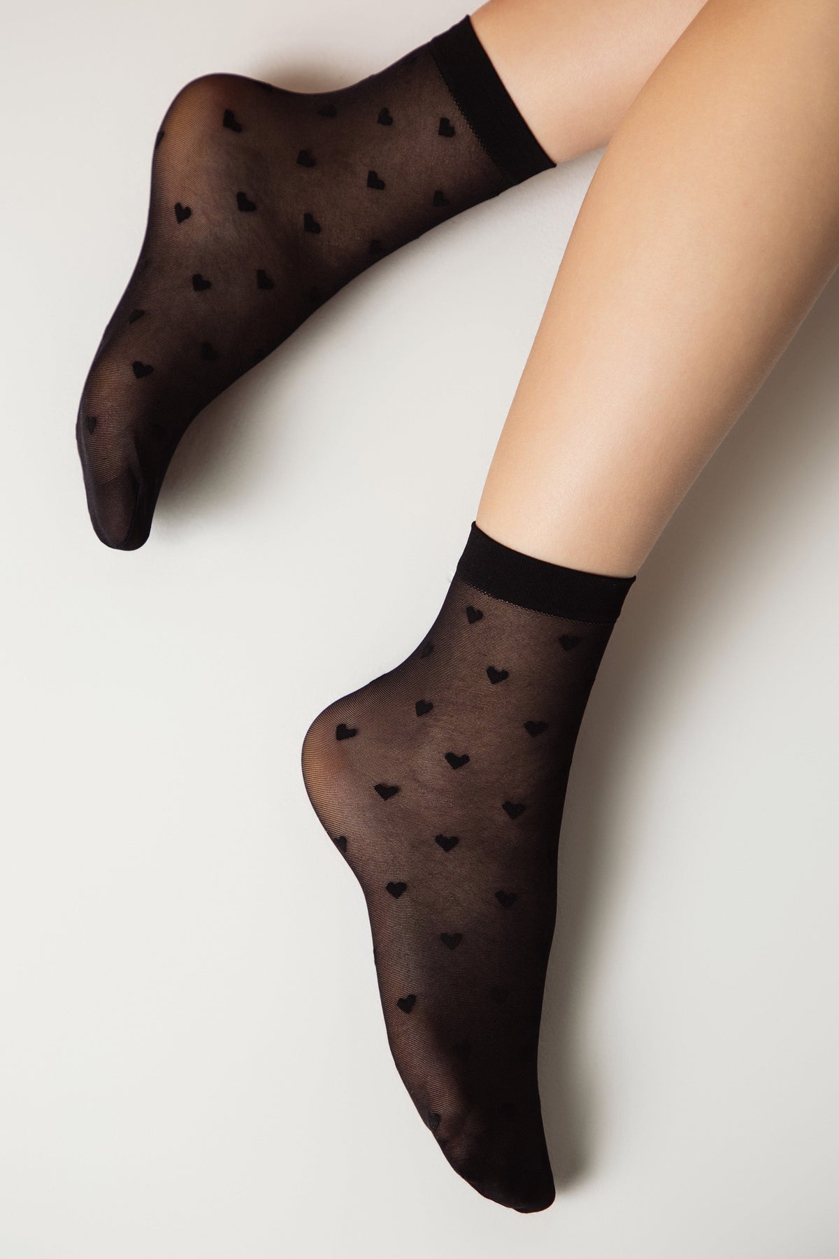 Sheer black ankle socks patterned with small black hearts worn on a person's feet against a plain white background.
