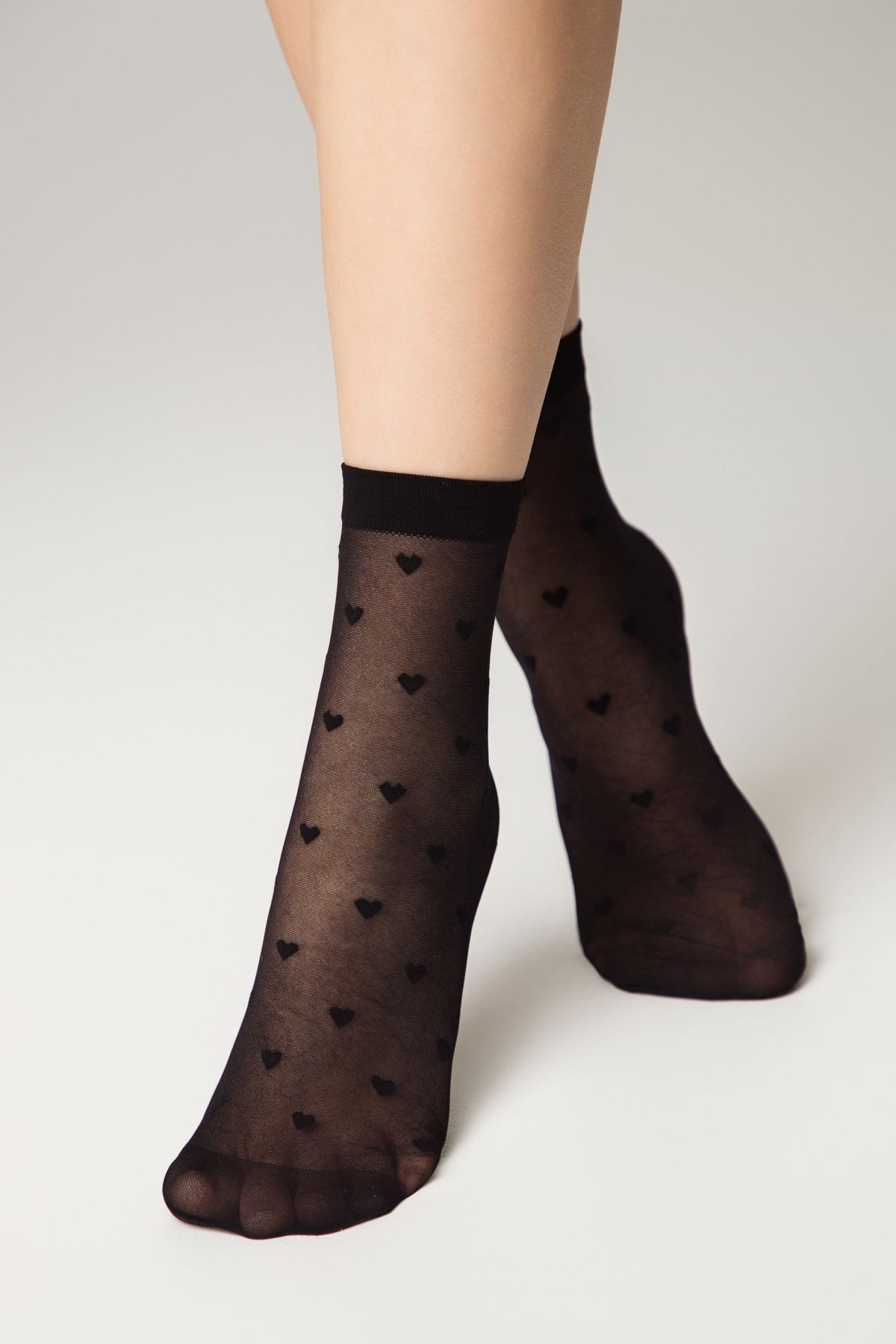 Sheer black ankle socks featuring a subtle, repeating pattern of small black hearts on a light grey background.