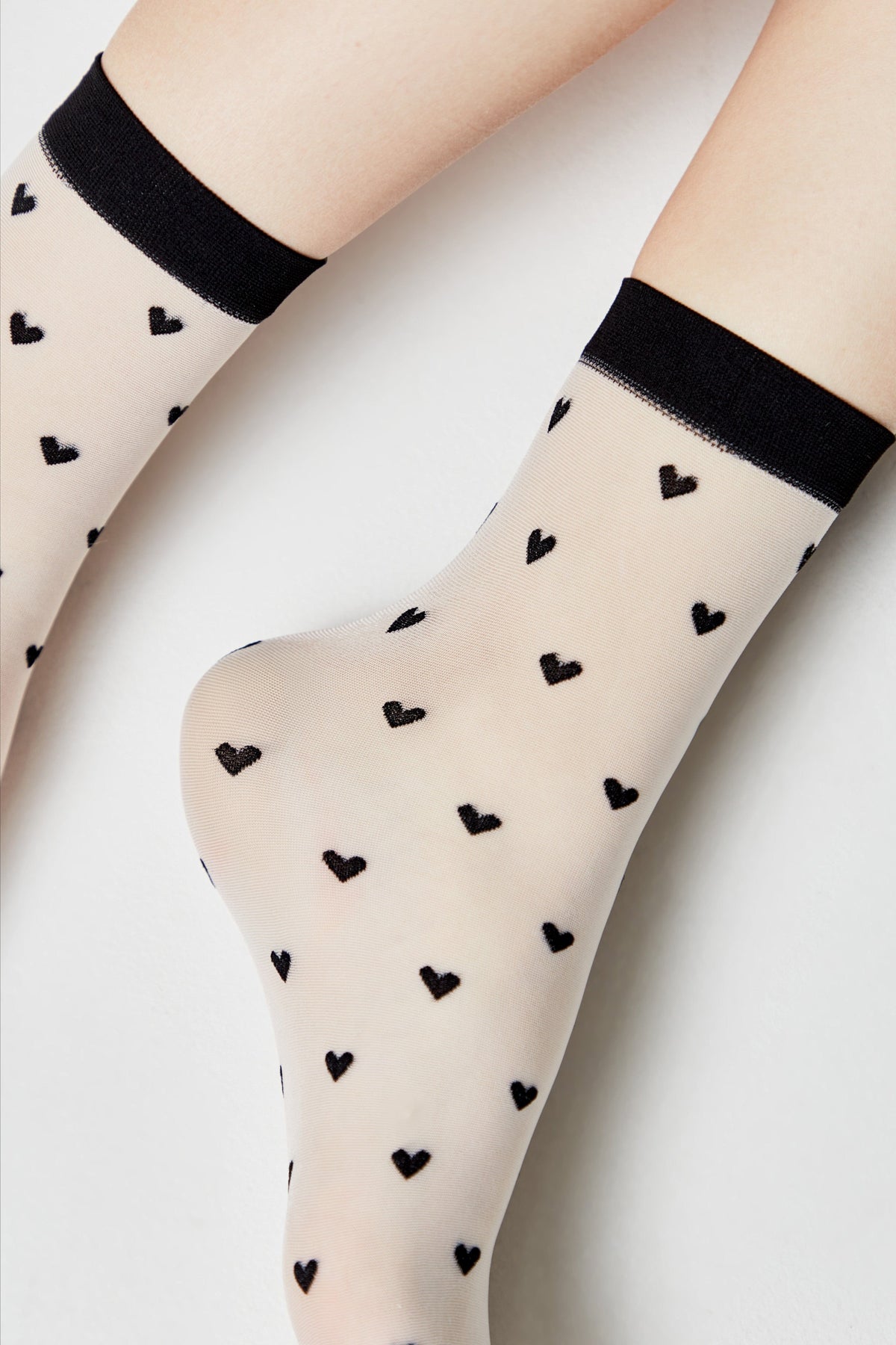 A pair of sheer, cream-colored ankle socks featuring a repeating pattern of small black hearts and finished with a solid black ribbed cuff.