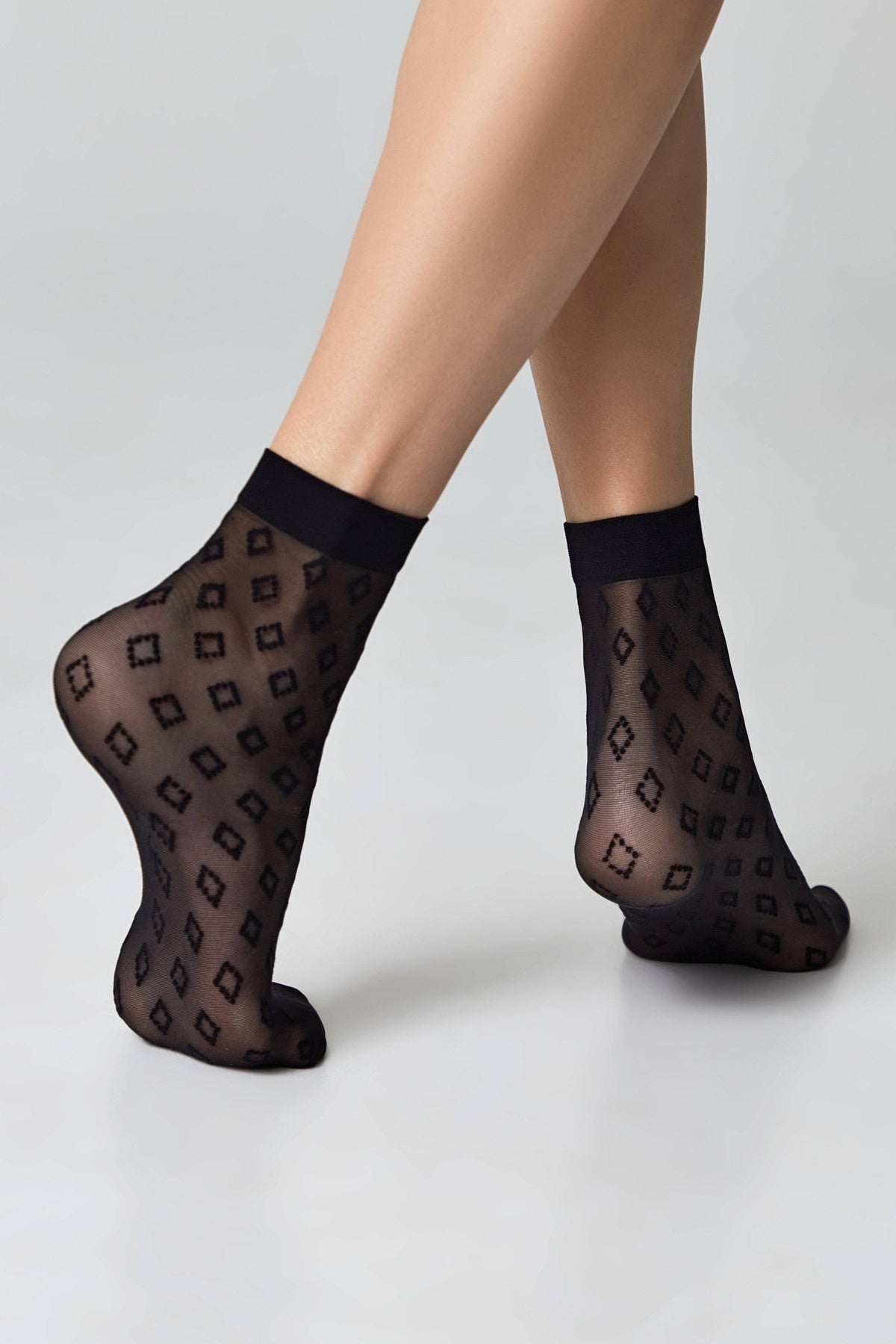 Sheer black ankle socks featuring a repeated geometric diamond pattern on a plain light grey background.