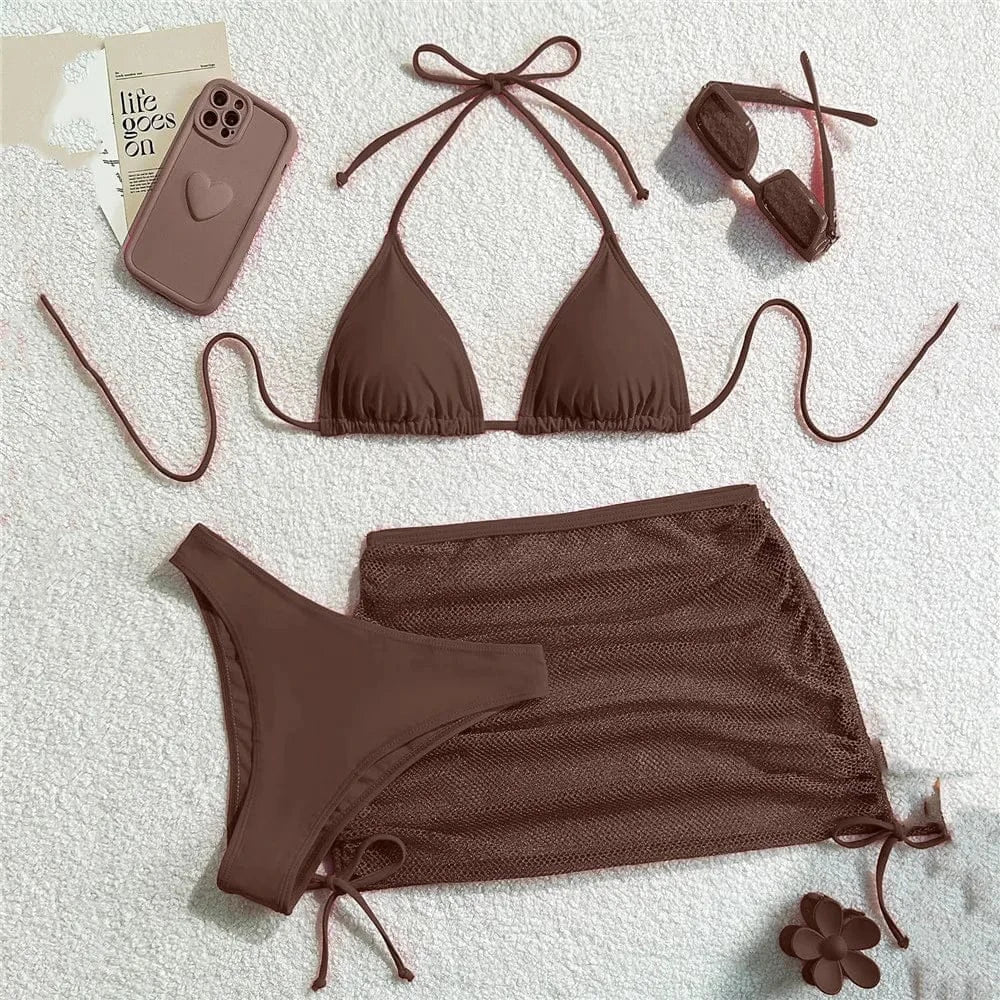 A brown three-piece bikini set consisting of a triangle top, a matching bikini bottom, and a sheer mesh skirt, displayed with a phone case, sunglasses, and a flower accessory on a white textured background.