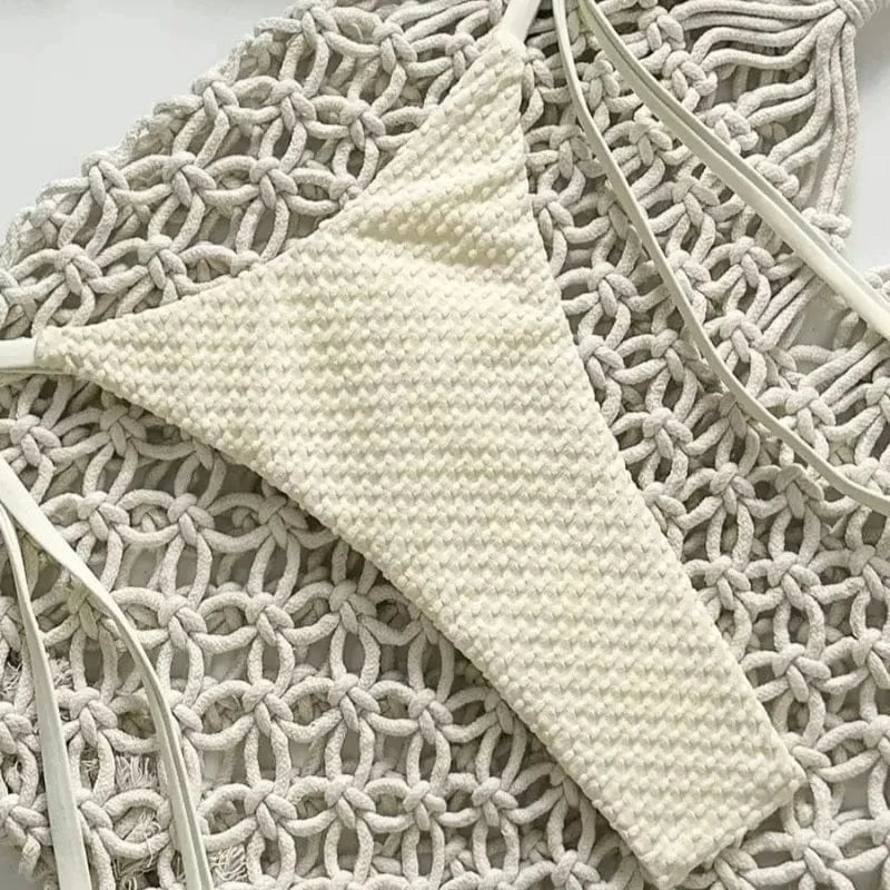 A cream-colored, textured bikini bottom with thin side straps rests on a decorative white macrame background.